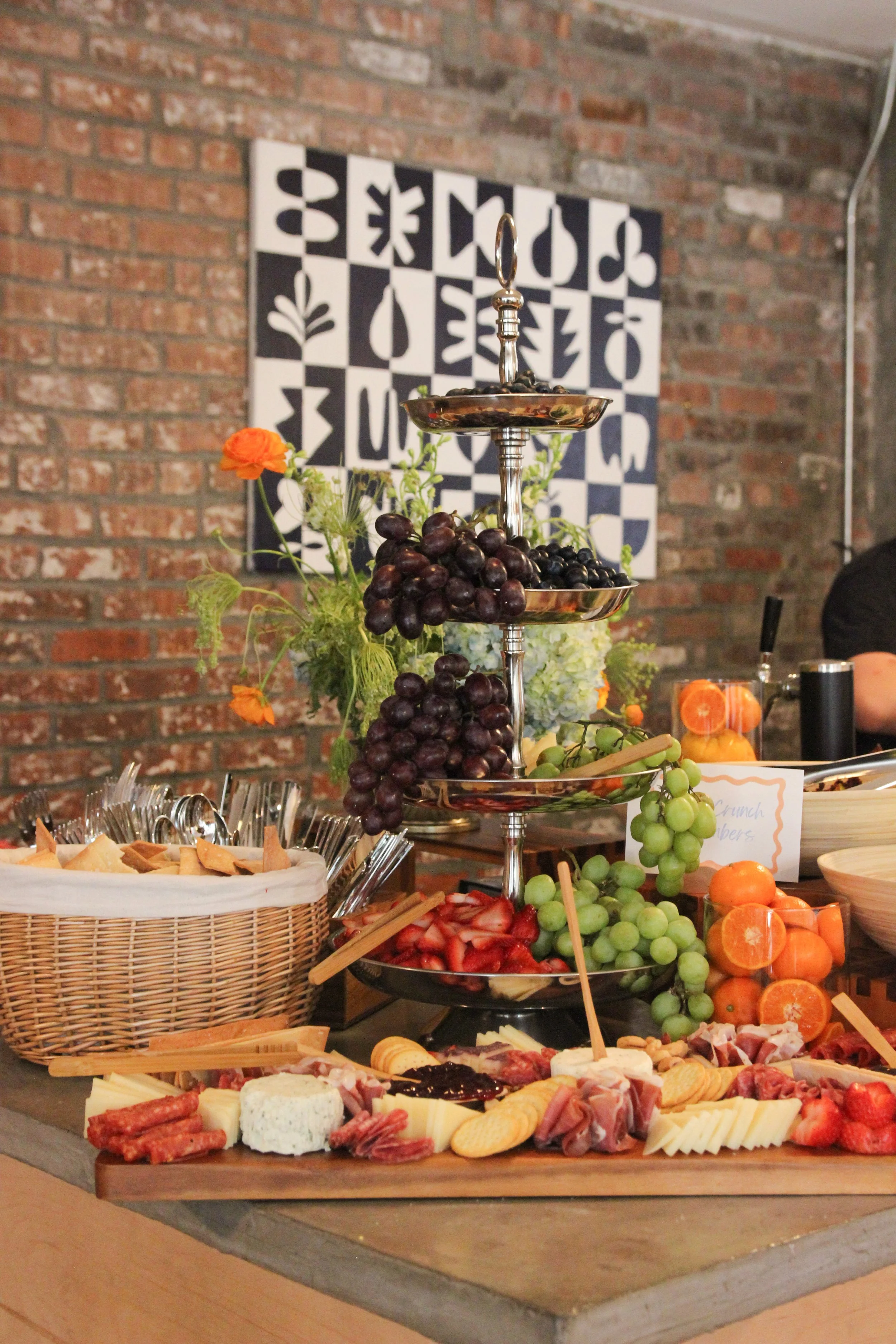 A buffet table with assorted cheeses, charcuterie, and fresh fruits including grapes, strawberries, and tangerines, set against a brick wall with black and white artwork.