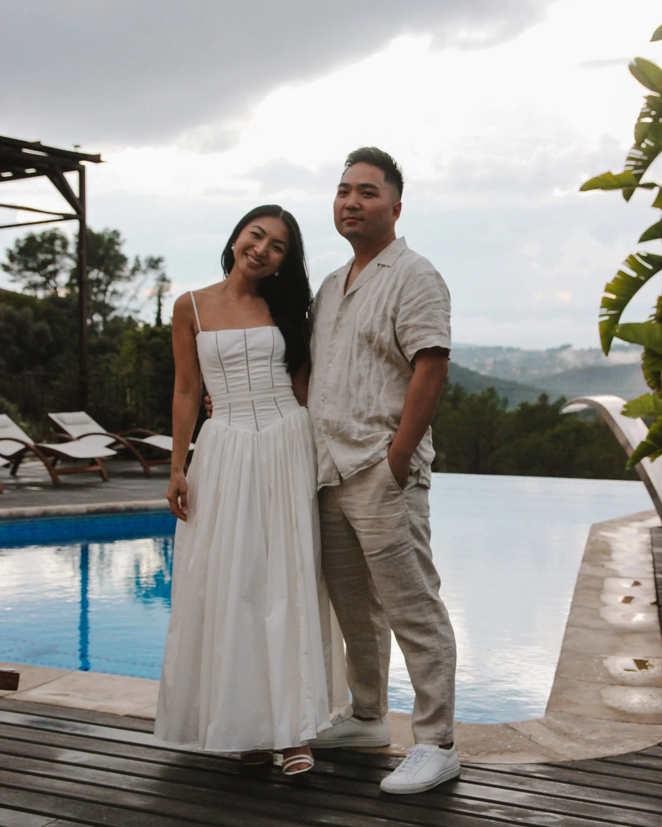 A smiling woman in a white dress and a man in light-colored casual clothing standing by a swimming pool with chairs, trees, and a cloudy sky in the background. The owners of Saturday Graze, a curated catering and grazing service.