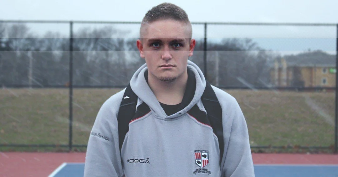 A young man with short hair wearing a gray hoodie and black backpack standing on an outdoor sports court.