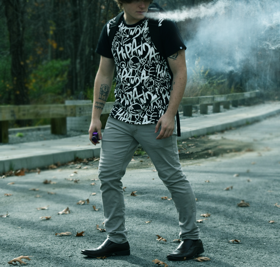 Person wearing a black graphic t-shirt and gray pants standing on a paved road scattered with fallen leaves, with trees and a wooden fence in the background.