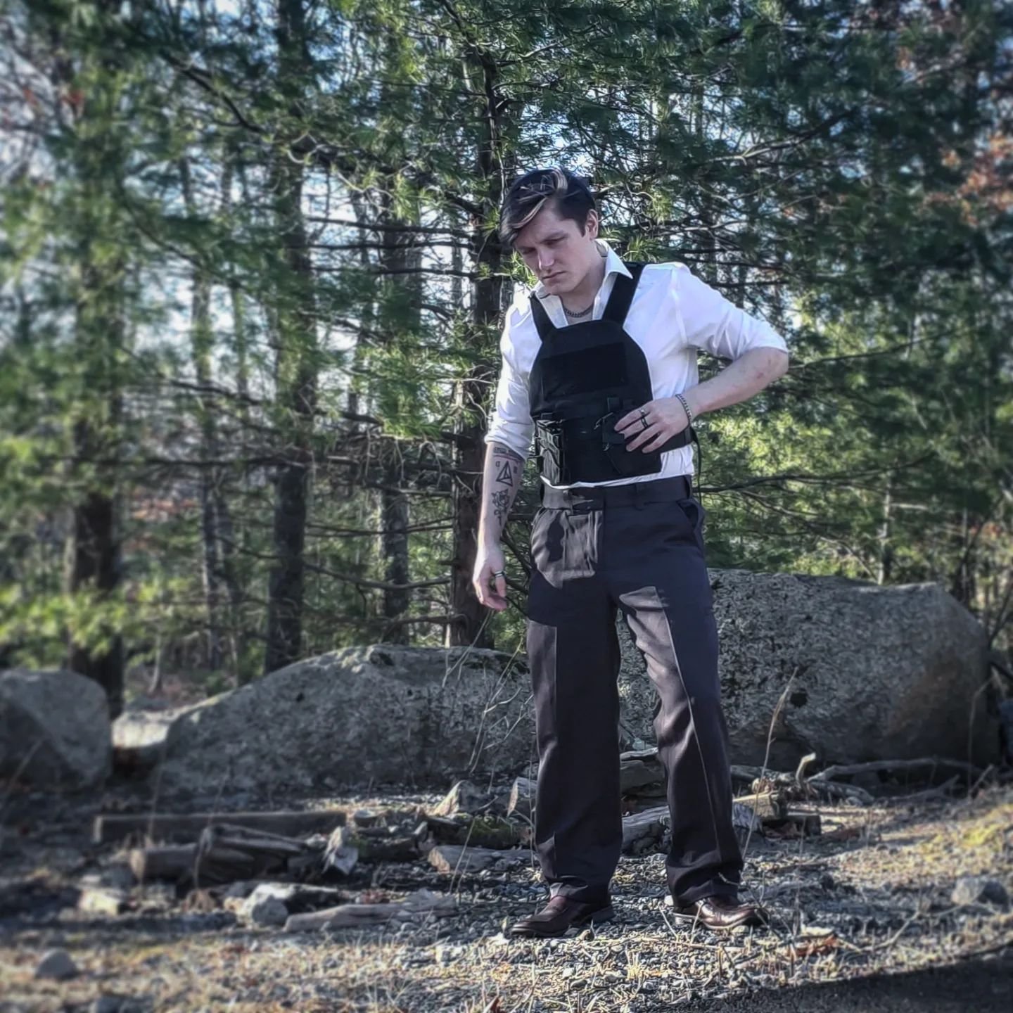 A young man with tattoos, wearing a white shirt and black pants, stands outdoors in a forested area with rocks and trees around him, looking down with a serious expression.