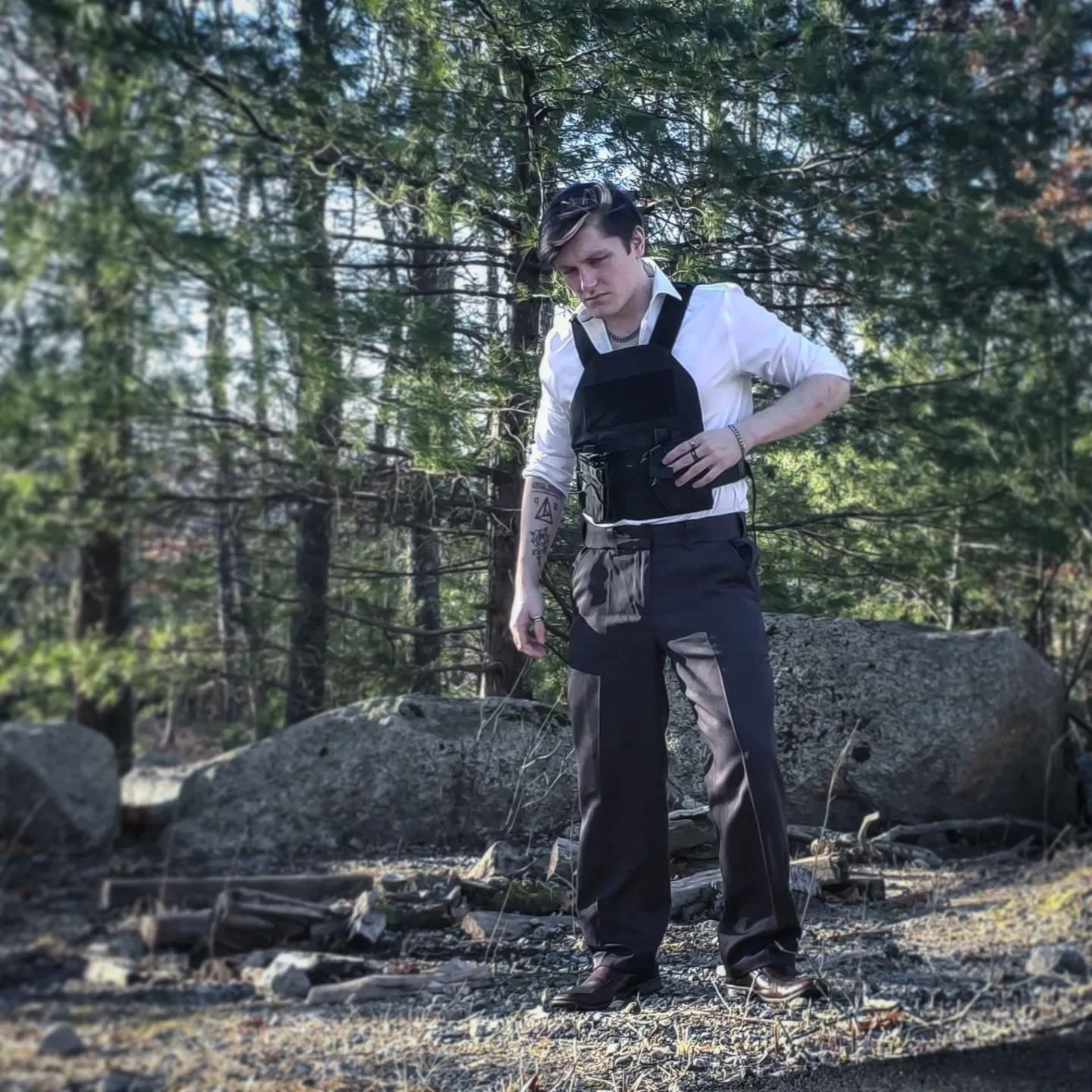 A young man with dark, styled hair, wearing a white shirt with rolled-up sleeves, black pants, and a small black backpack, stands outdoors on rocky terrain surrounded by trees. He appears to be looking down at the ground with a serious expression.