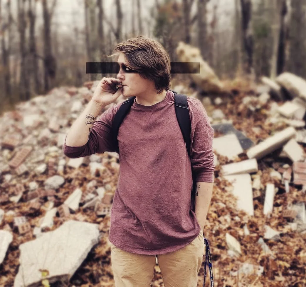 Person with short brown hair, black face mask, wearing a maroon long-sleeve shirt, standing outdoors in a wooded area with fallen leaves and scattered debris.