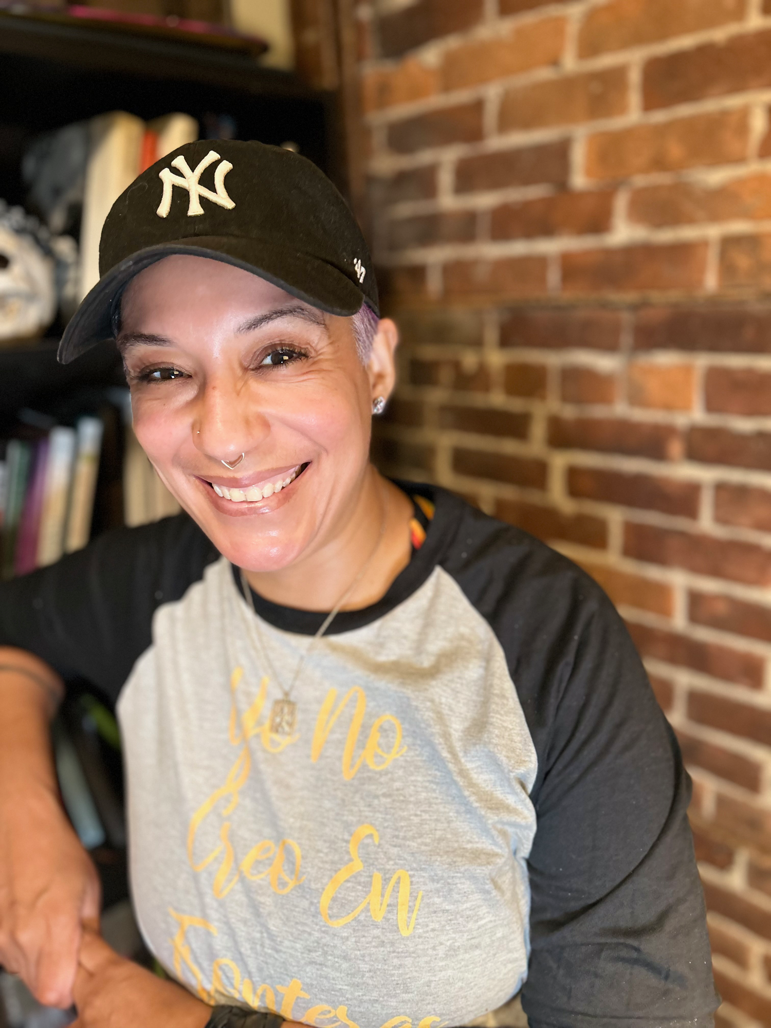 A smiling woman with short hair wearing a black New York Yankees cap and earrings, standing in front of a brick wall.