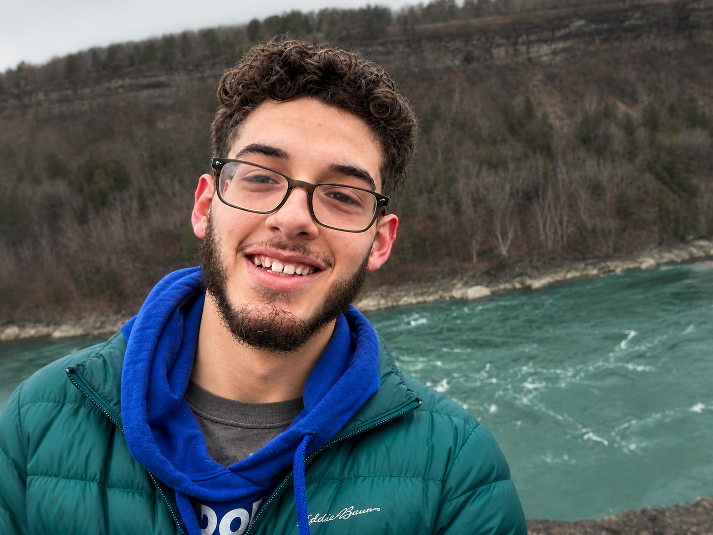 Young man with glasses and a beard smiling in front of a river and hillside with trees.