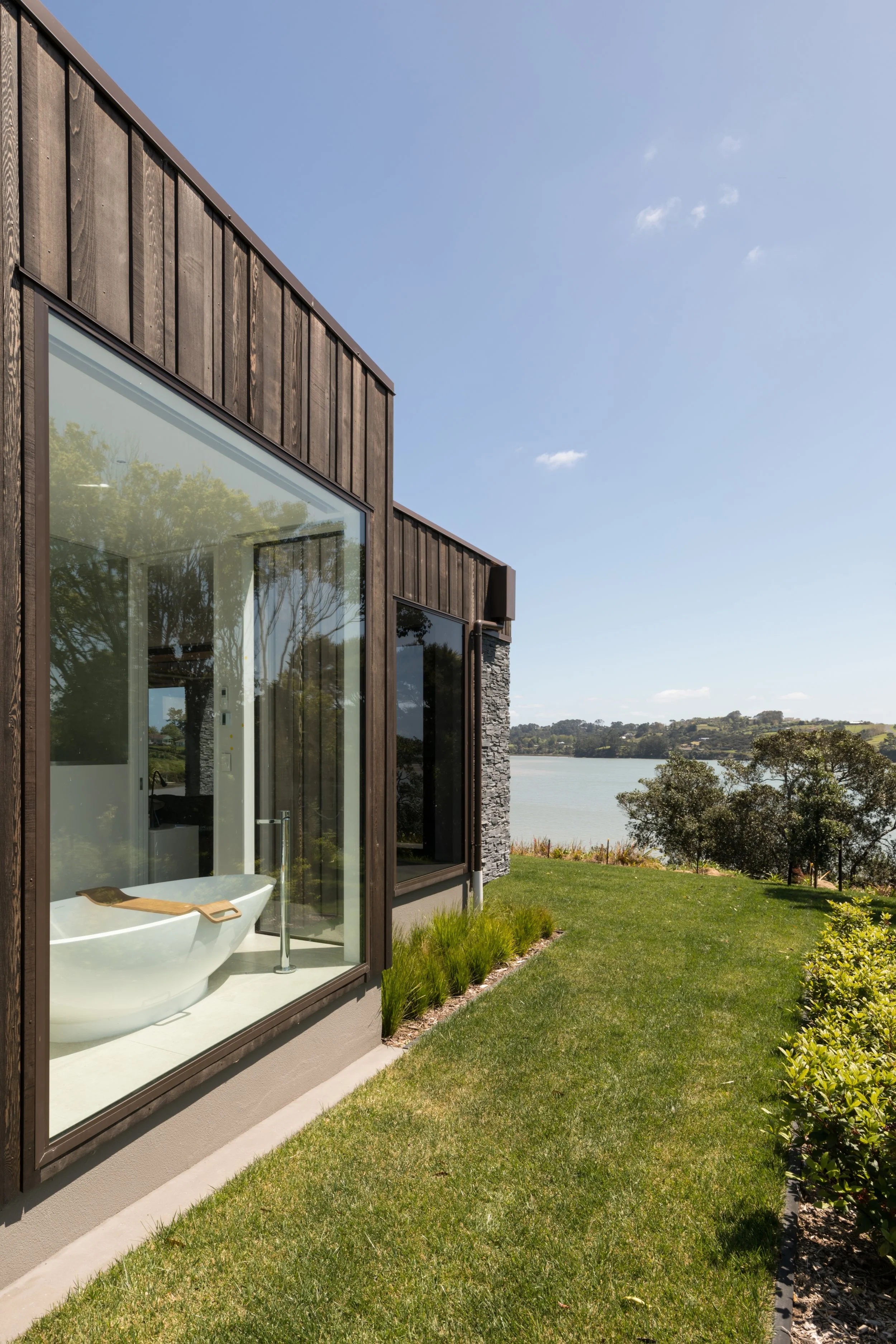 Custom architectural modern home with large glass windows overlooking a river, surrounded by green grass and trees on a sunny day.
