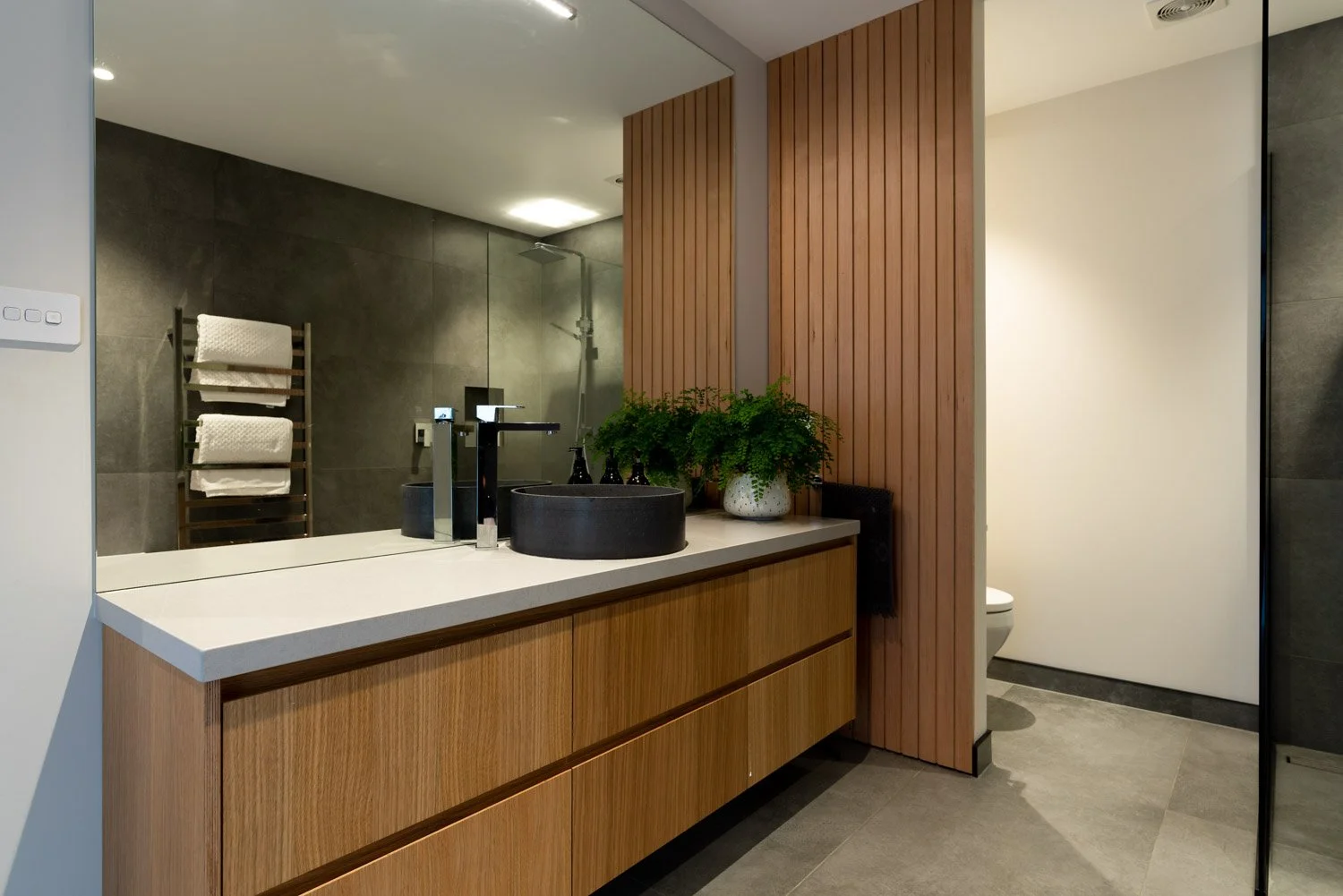 Modern bathroom with wooden vanity, black vessel sink, and a large mirror. To the right, a partial view of a white toilet, with wood-paneled wall and gray tiled floor. Towel rack with white towels is reflected in the mirror.