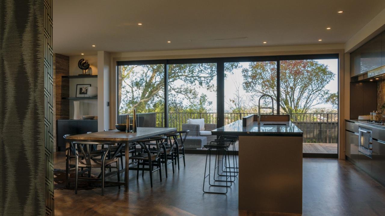 Kitchen with large sliding glass doors opening to a balcony with trees outside. Dining table with chairs, kitchen island with stools, and modern appliances.