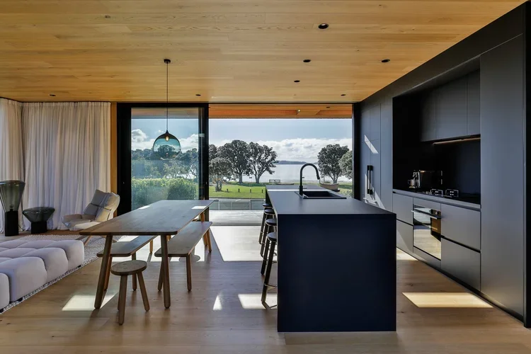 Modern custom open-concept kitchen and dining area with large stunning windows overlooking the beach featuring a wooden ceiling. 