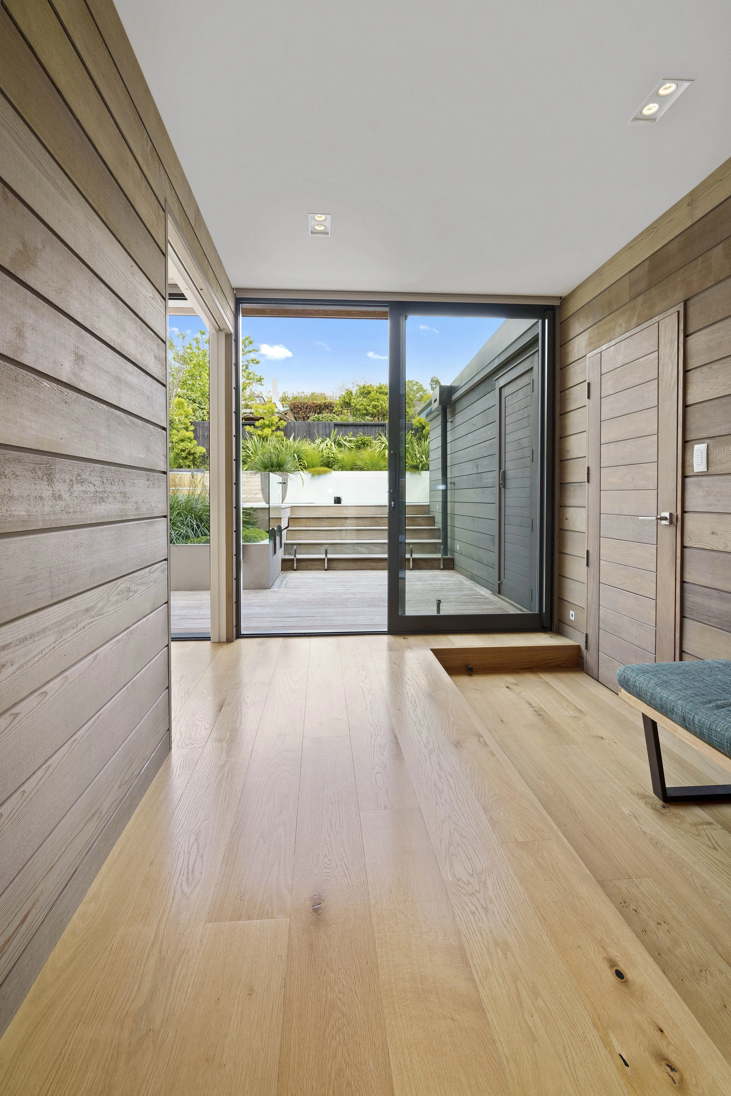 Interior view of a room with wooden walls and floor, glass sliding doors leading outside to a garden with plants and stairs, and partial view of a chair with teal cushion.