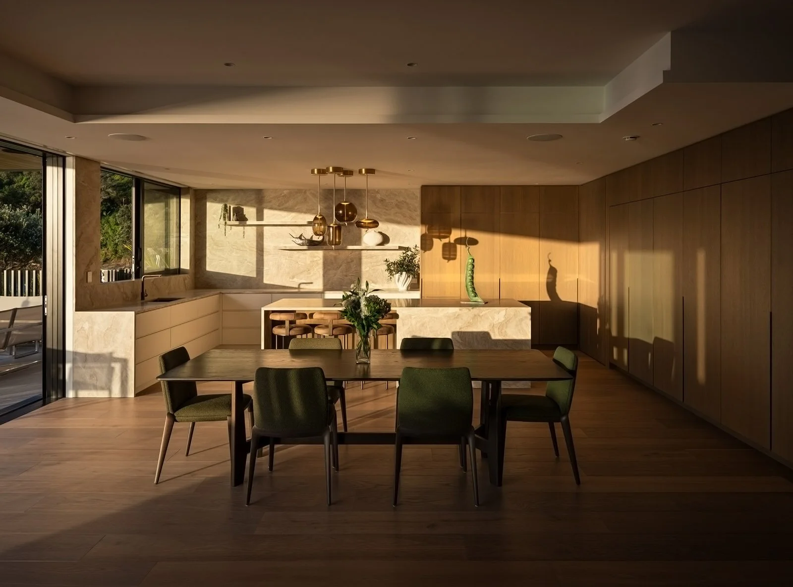 Golden hour, Modern dining area with wooden table, six green chairs, a vase with flowers, and sunlight cast shadows on the walls and floor. The Kitchen features stunning Taj Mahal Stone spanning up to the ceiling and custom cabinetry.