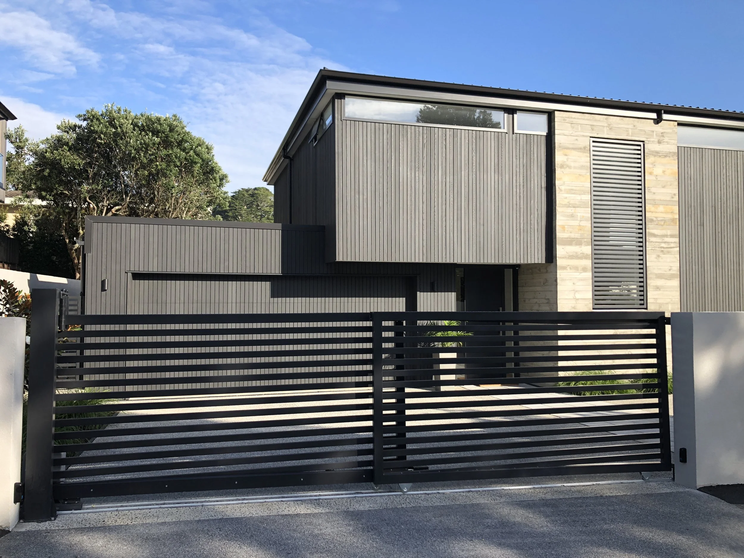 Modern house with a black gate in the foreground, wood and metal exterior, and a large window near the roof. Trees and blue sky with clouds are visible in the background.