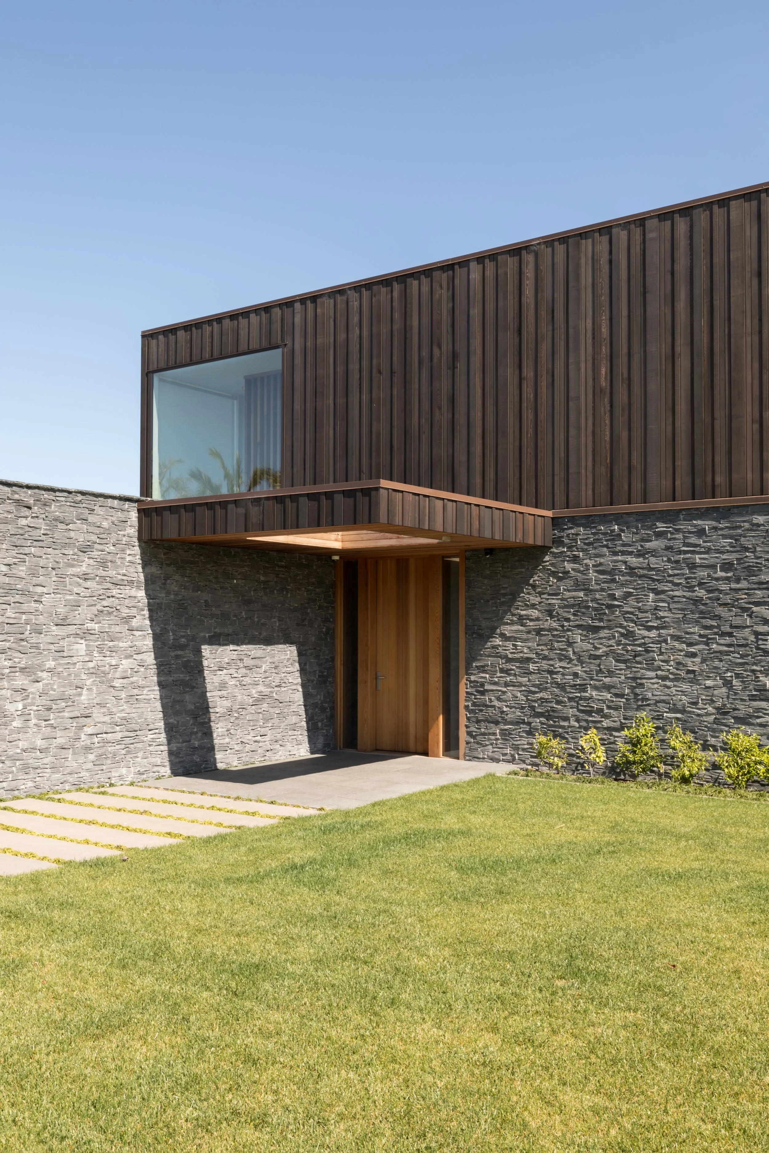 Modern house exterior with stone wall, dark wooden paneling, and a small balcony with trees visible through the window. Well-manicured lawn and a concrete pathway leading to the wooden front door.