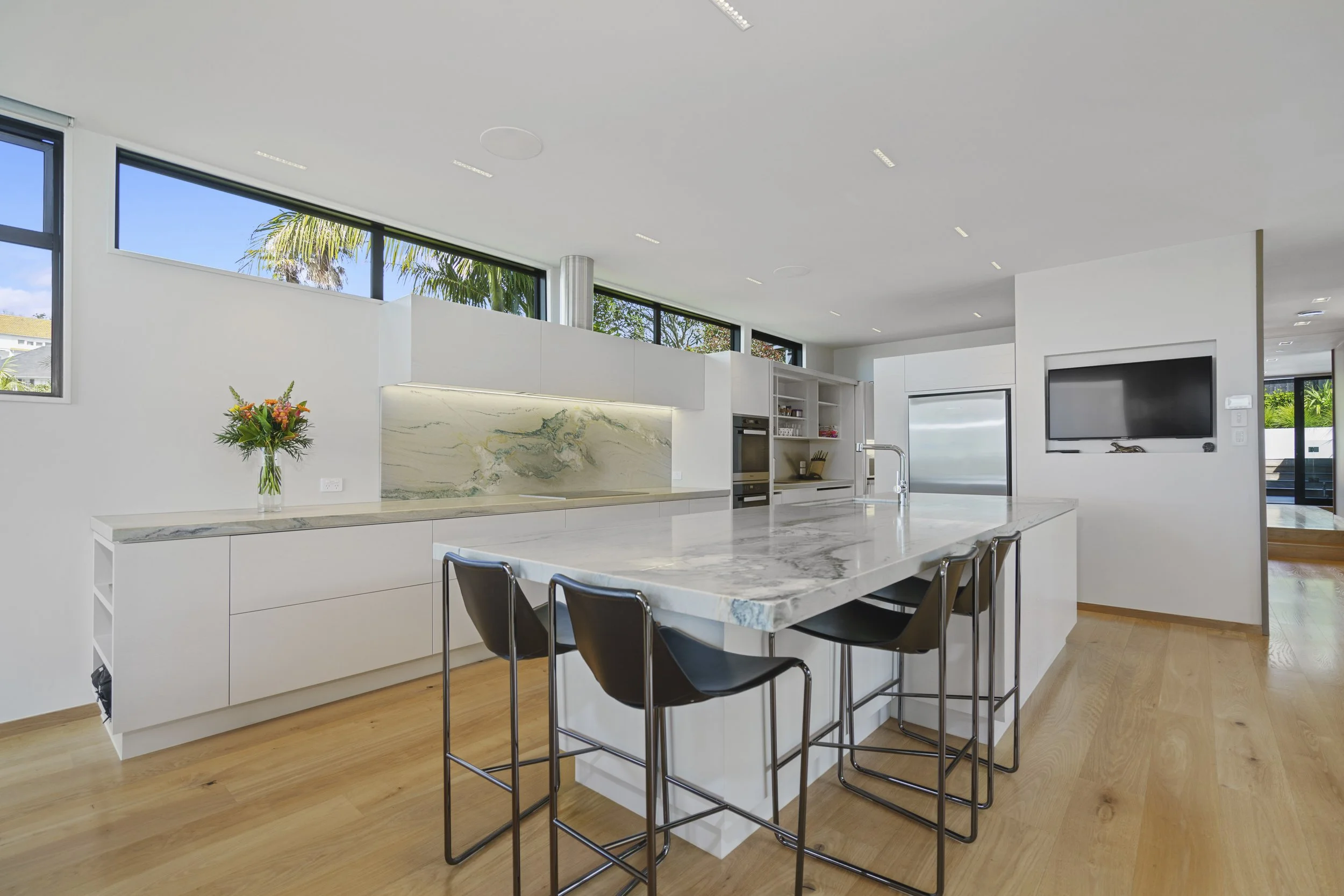 Modern kitchen with white cabinets, marble island, black chairs, large windows, and a flat-screen TV on the wall.