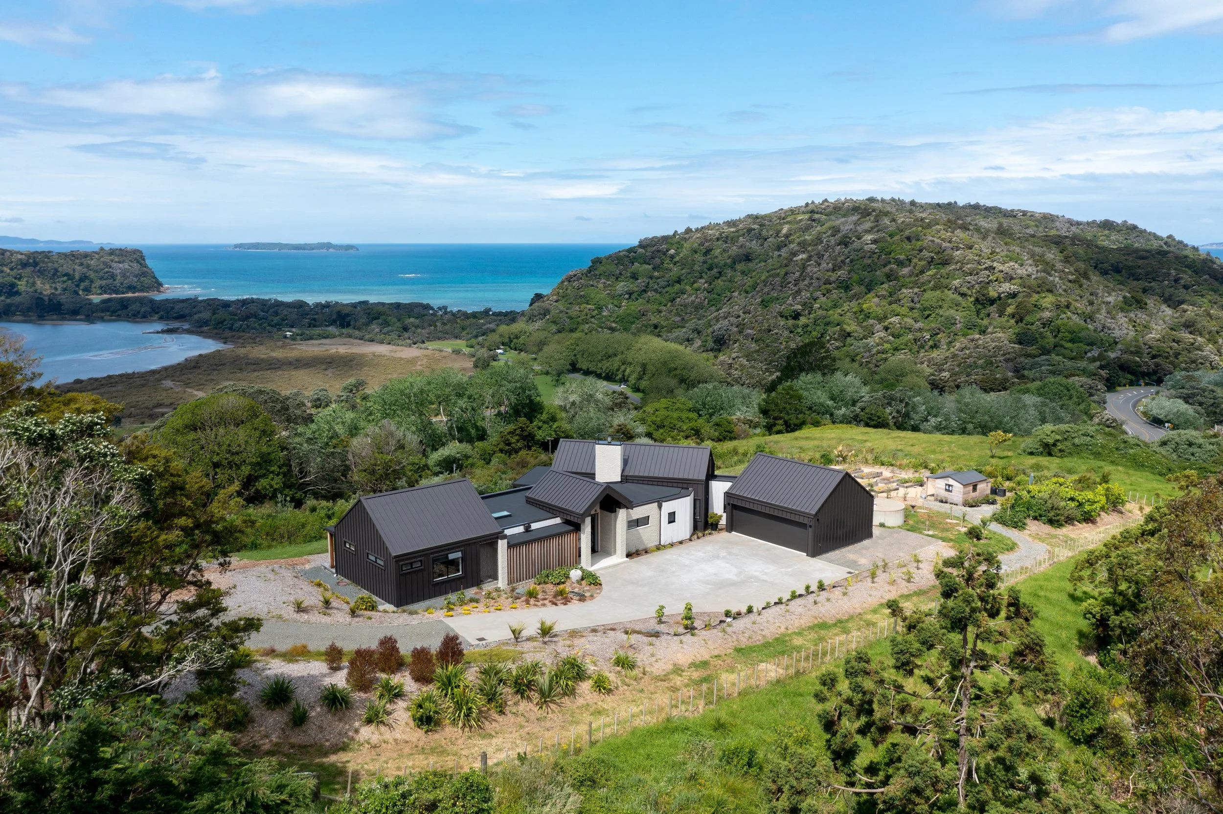 A panoramic view of a coastal landscape with modern architectural custom home in the foreground, surrounded by lush green hills leading to the ocean, with a partly cloudy sky overhead.