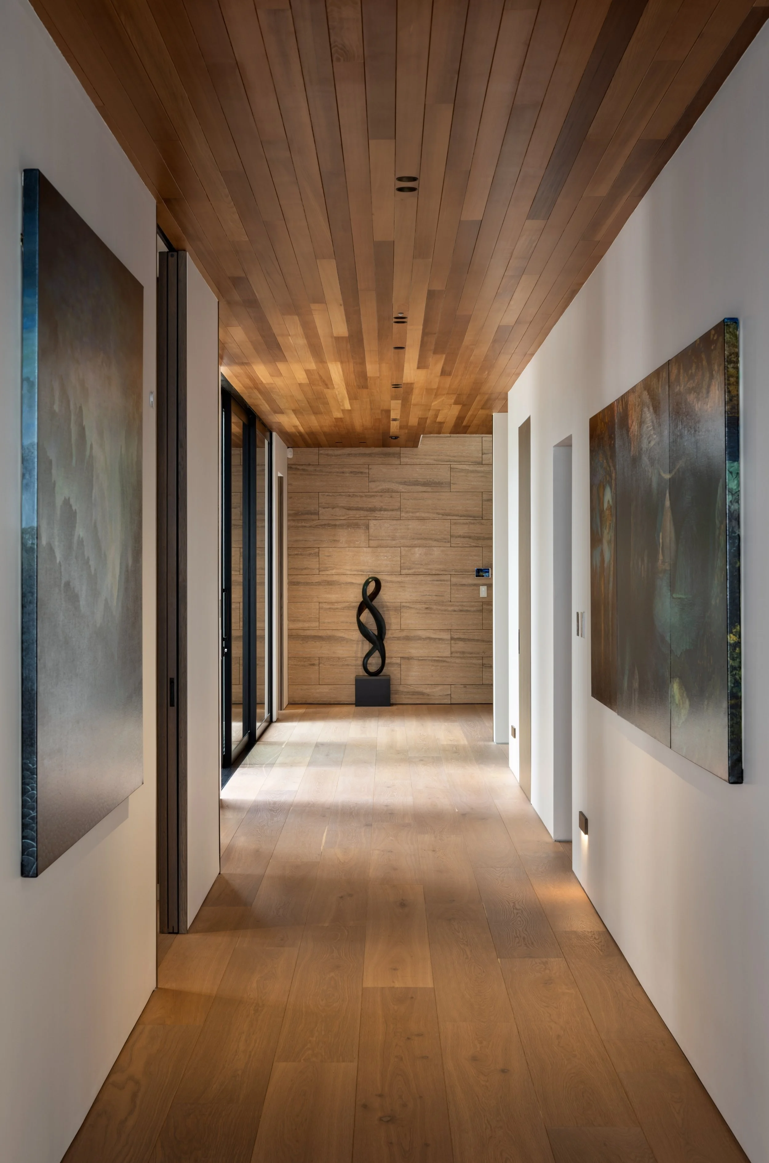 A high-end architectural hallway with wooden flooring and a feature cedar wood ceiling, featuring artwork on the walls and a sculpture at the end. Featuring high quality materials and quality workmanship. 