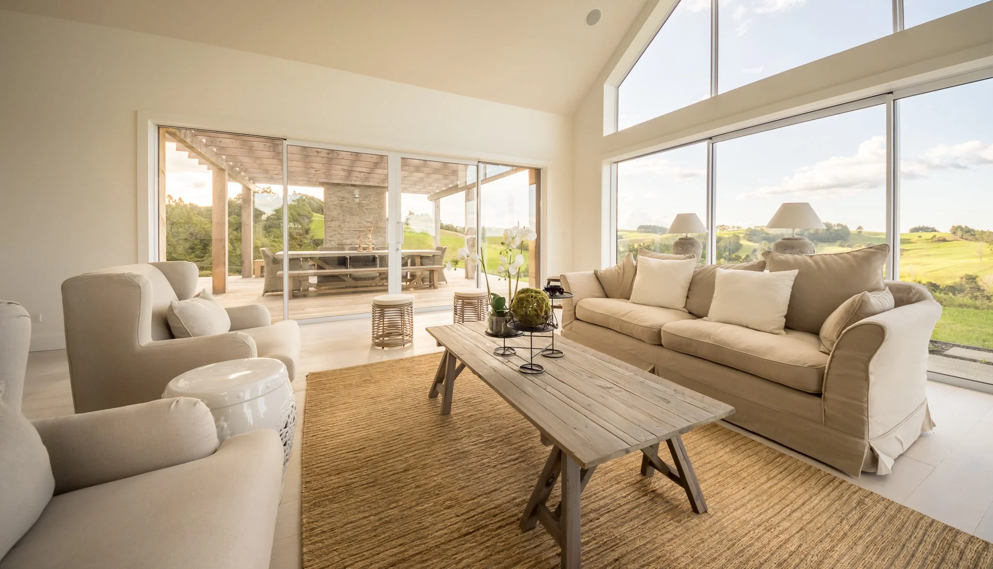 Bright living room with large glass windows and sliding doors showing a green outdoor landscape, featuring a beige sofa with cushions, patterned armchairs, wooden coffee table with plants, and a view of the outdoor patio with a dining table.