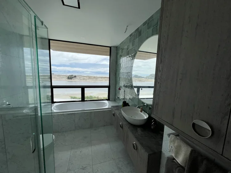 Modern bathroom with a large window overlooking a body of water and distant hills, featuring a bathtub, a vessel sink, and a glass shower enclosure.