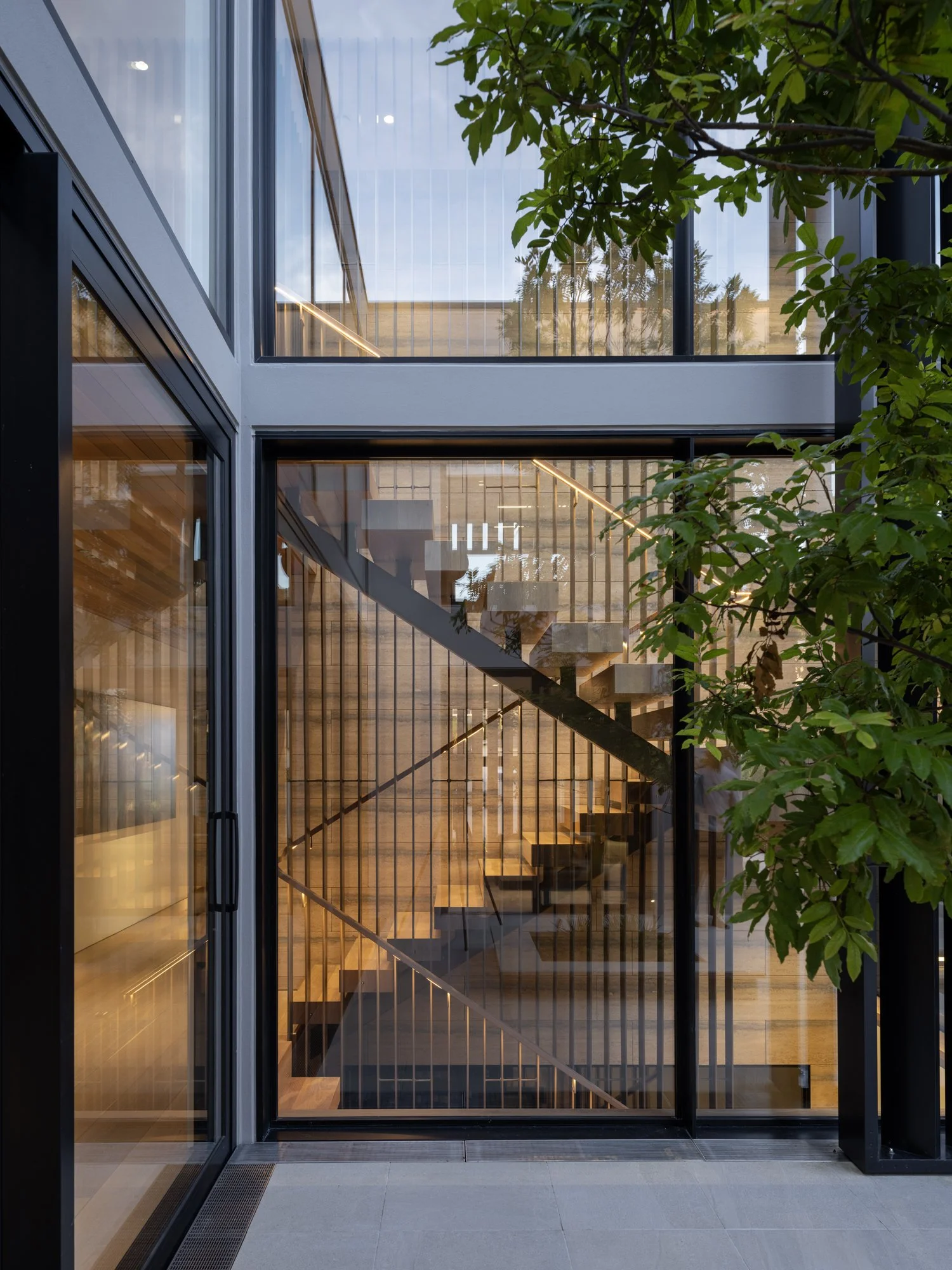 View through glass doors revealing a modern custom brass coated 8.5 meter staircase feature in the heart of the home, sur by stunning wood floating stairs and travertine tile. Highlighted through large windows and light from the courtyard.