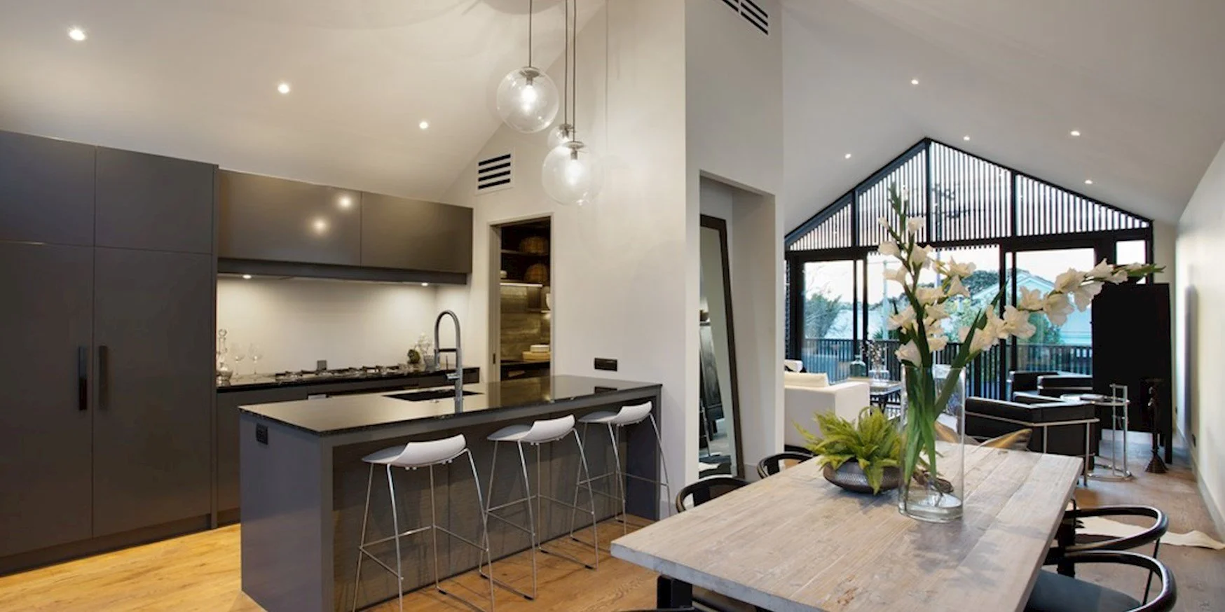 Open-plan kitchen and living area with modern design, featuring a kitchen island, white bar stools, dark cabinetry, a wooden dining table with a vase of white flowers, and a sitting area with large windows, black furniture, and outdoor views.