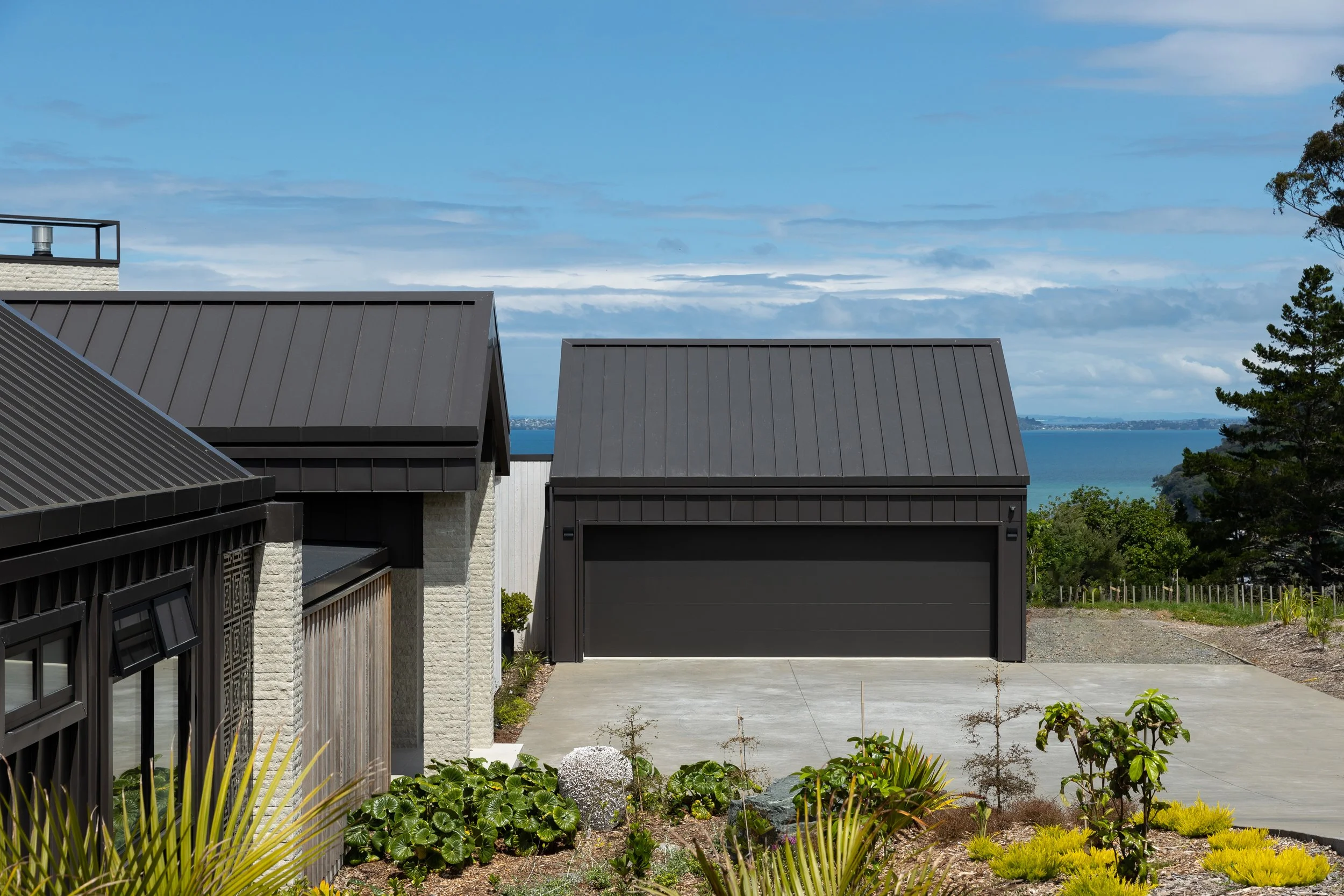 View of modern house with black metal roof and black garage door, overlooking water, with landscaping plants in foreground and trees in background.