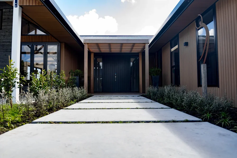 Modern house entrance with a pathway leading to a black front door, surrounded by landscaped plants and shrubs, with glass windows and wooden siding.
