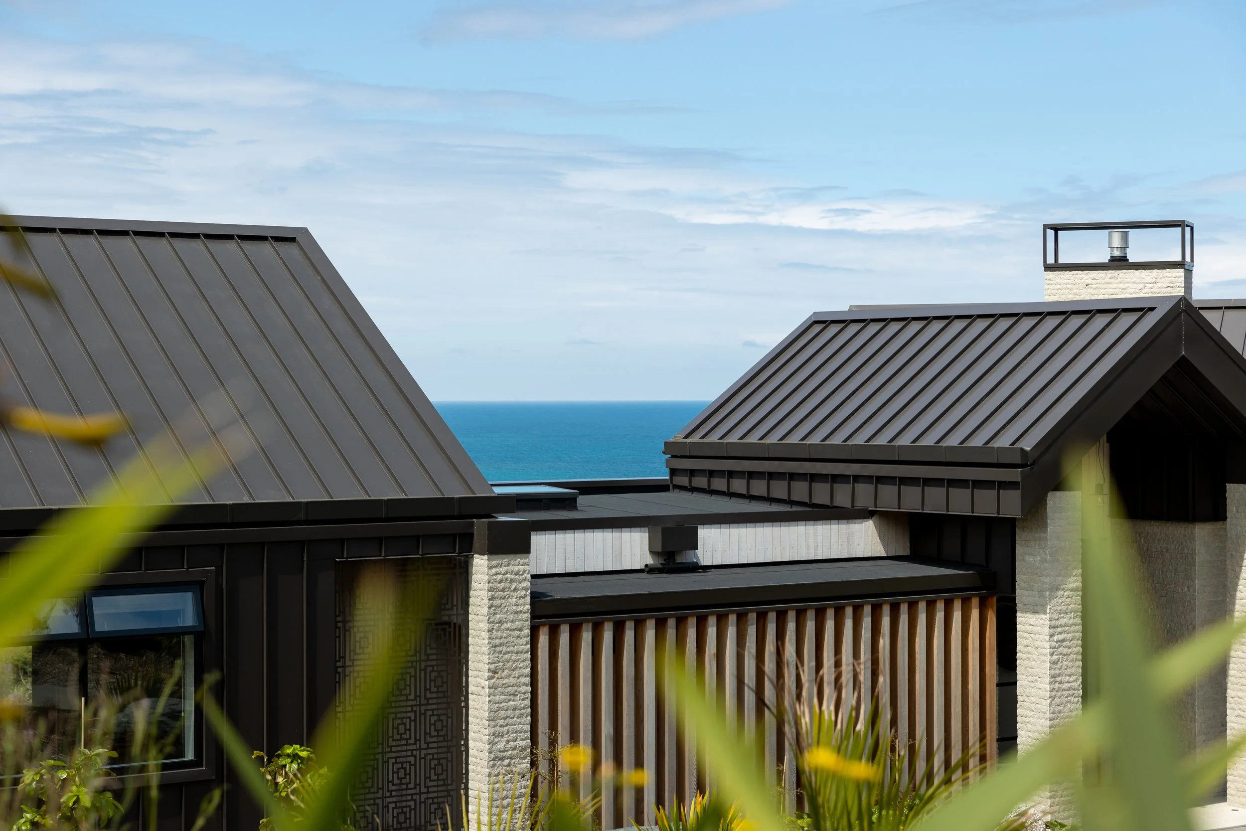A view of a custom architectural modern house rooftops with black metal roofing, a chimney with a light, and a glimpse of the ocean in the background under a partly cloudy sky.