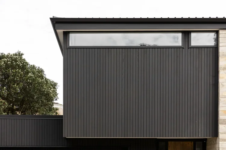 Close-up of a modern building's exterior with dark vertical siding, a horizontal window near the roof, and a small section of lighter-colored stone wall on the right.