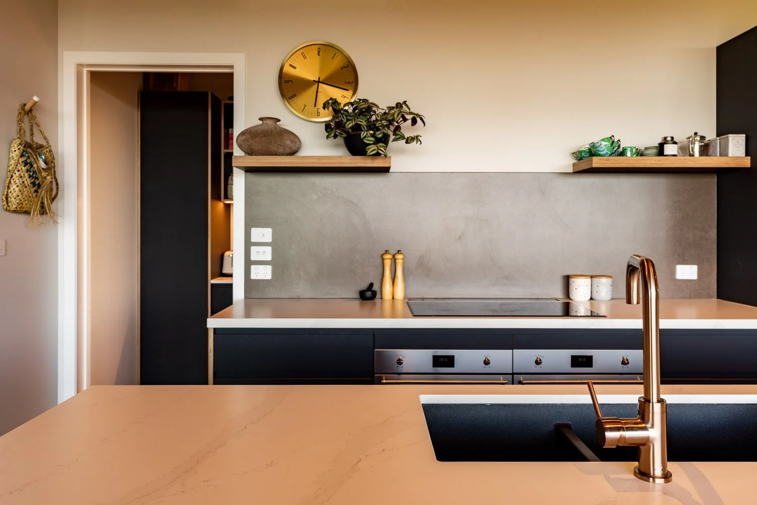 Modern kitchen with beige countertop, gold faucet, dark cabinets, wall-mounted wooden shelves, potted plants, clock, and kitchen accessories.