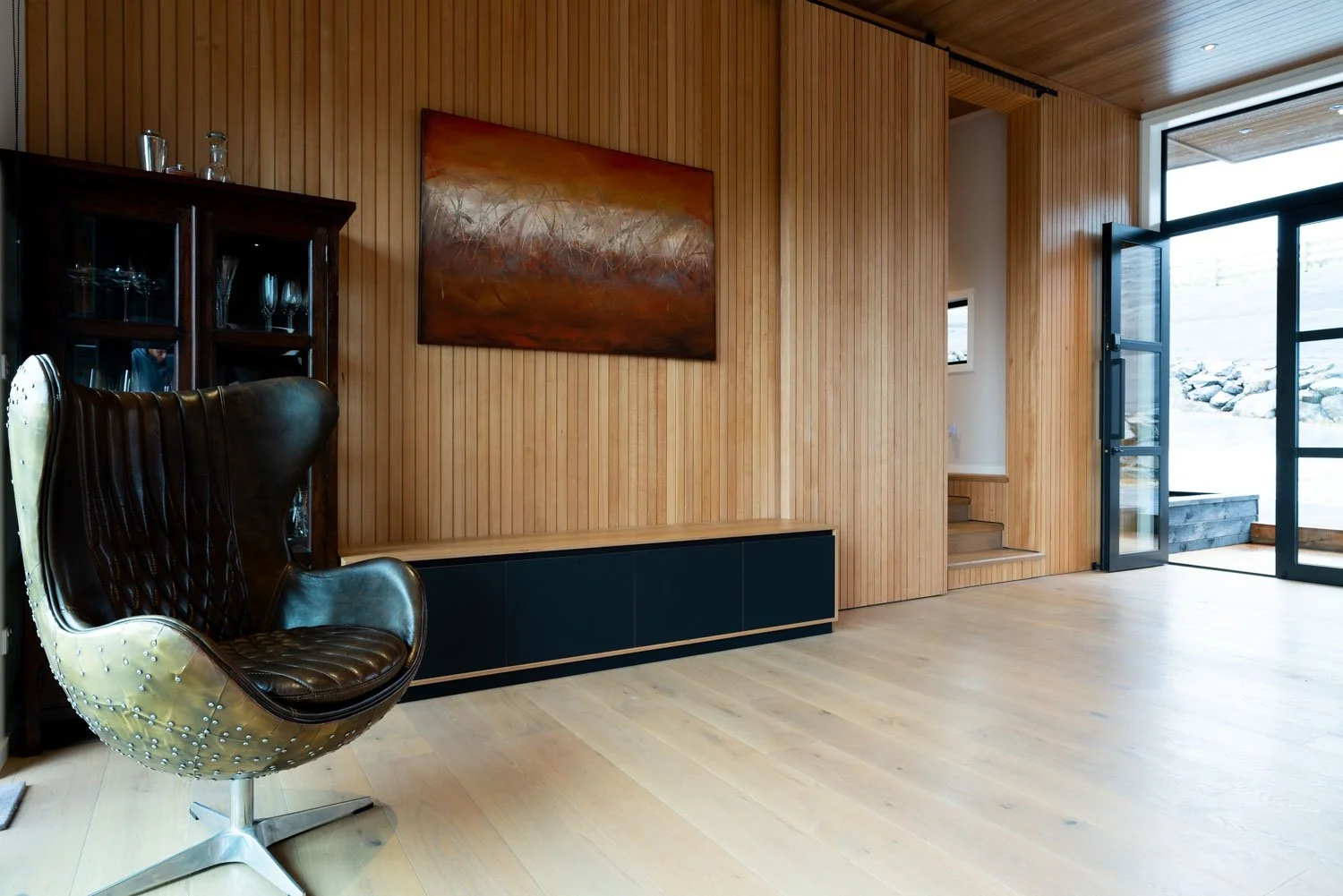 Interior of a modern home with wooden panel walls, a black cabinet, a mounted painting with warm tones, a leather and metal chair, and large glass doors opening to a snowy outdoor landscape.