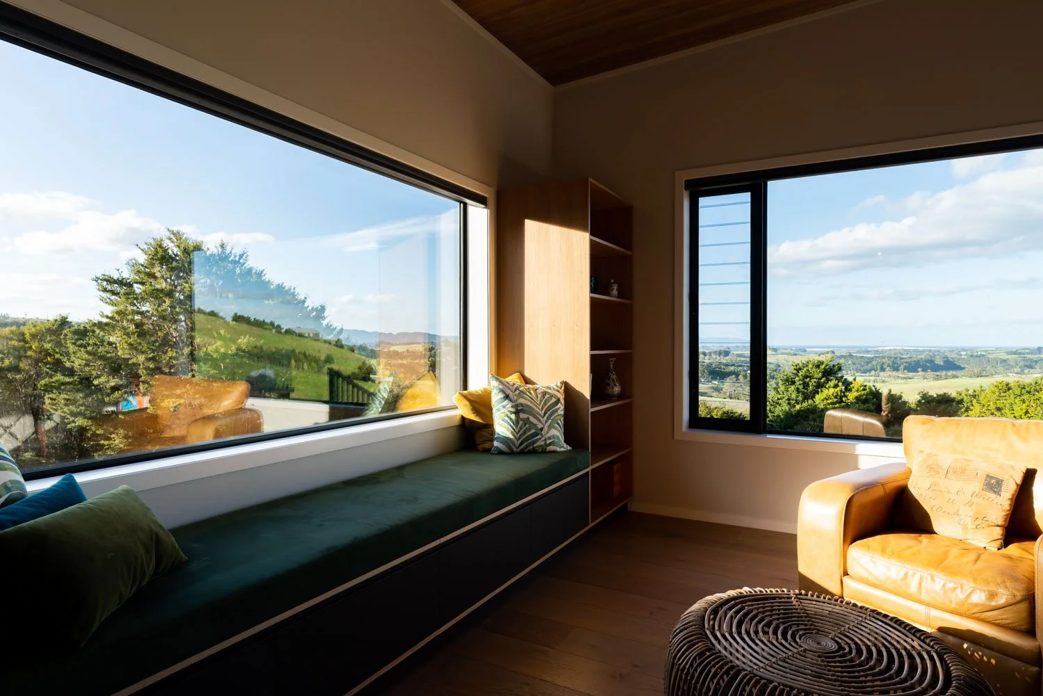 A cozy living room with large windows showing a scenic landscape of trees and hills, a green window seat with decorative pillows, and a leather armchair with a pillow near a circular woven table.