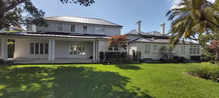 A large white house with a metal roof, surrounded by a green lawn and trees including palm trees. The house has multiple windows and a covered porch.