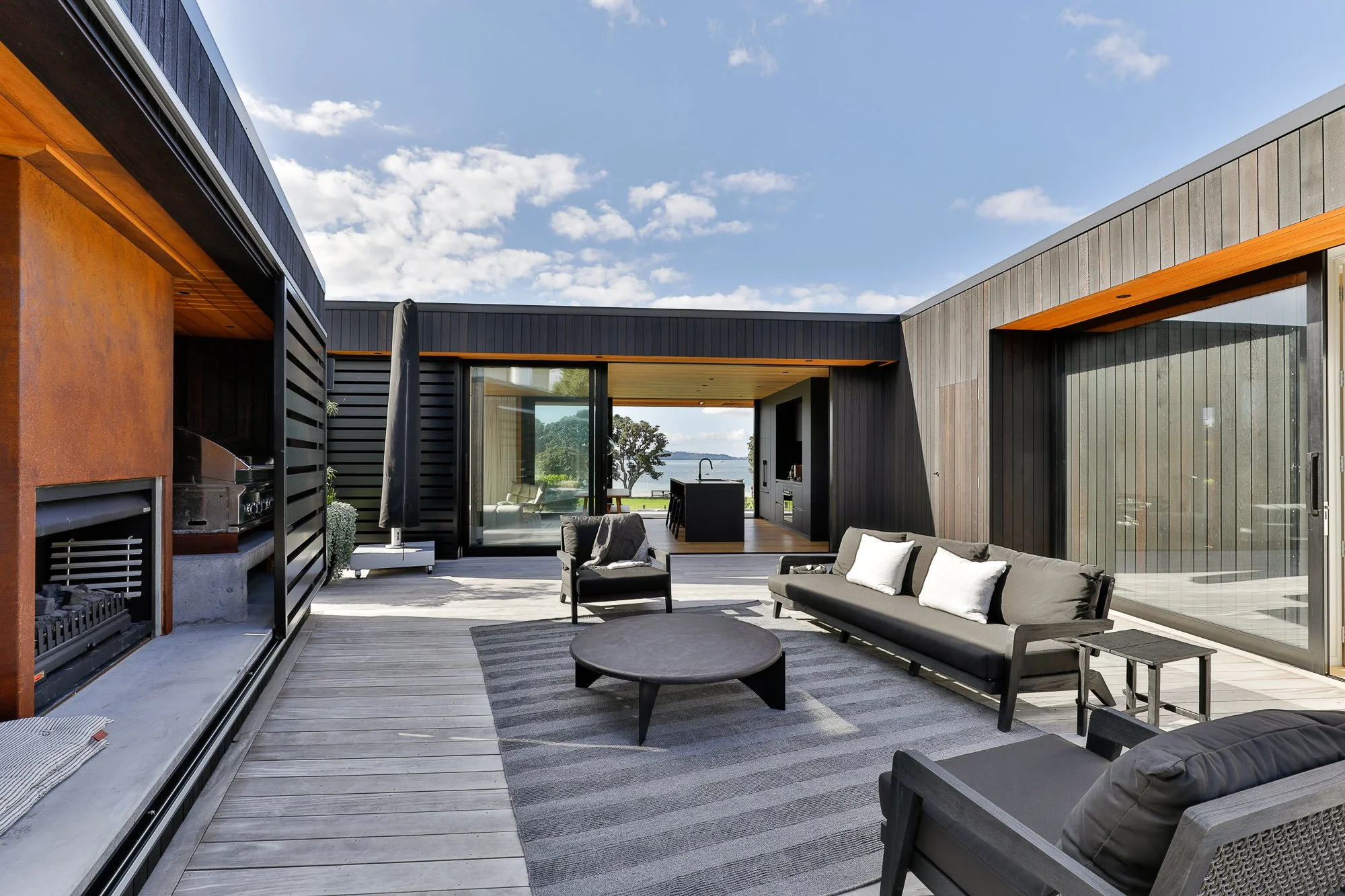  Custom outdoor patio with black and gray furniture, including sofas and chairs with white pillows, on a wooden deck with an outdoor BBQ area and large sliding glass doors leading to indoor living space, overlooking a water view.