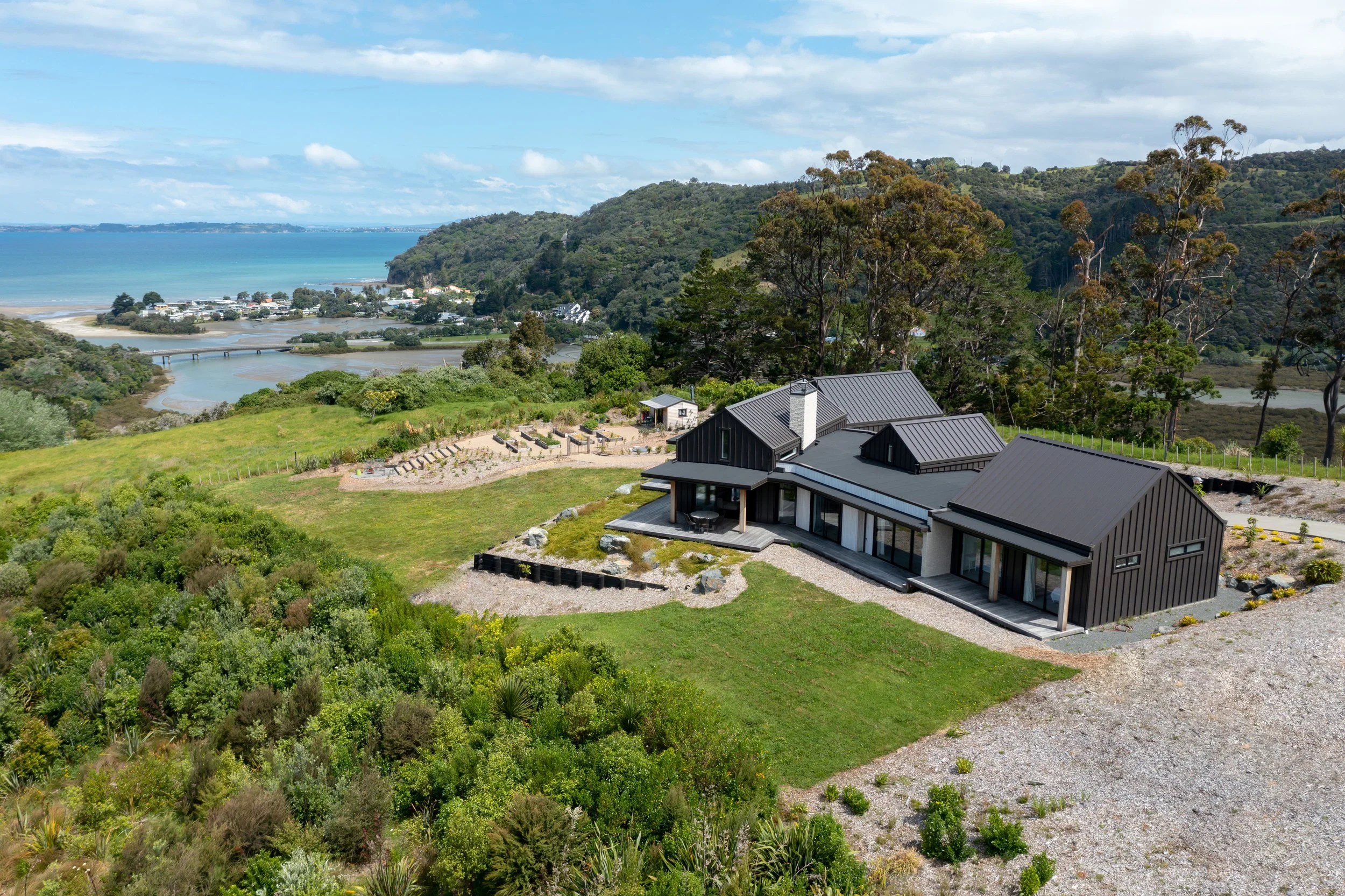 Modern house with dark exterior sitting on a grassy hillside overlooking a river and town, with dense forest and hills in the background on a partly cloudy day.