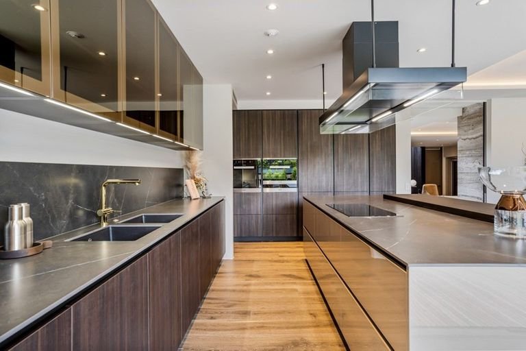 Modern kitchen with dark wood cabinets, black marble countertops, and light hardwood floors. Features a double sink, built-in oven, and a large island with a cooktop and a suspended range hood.