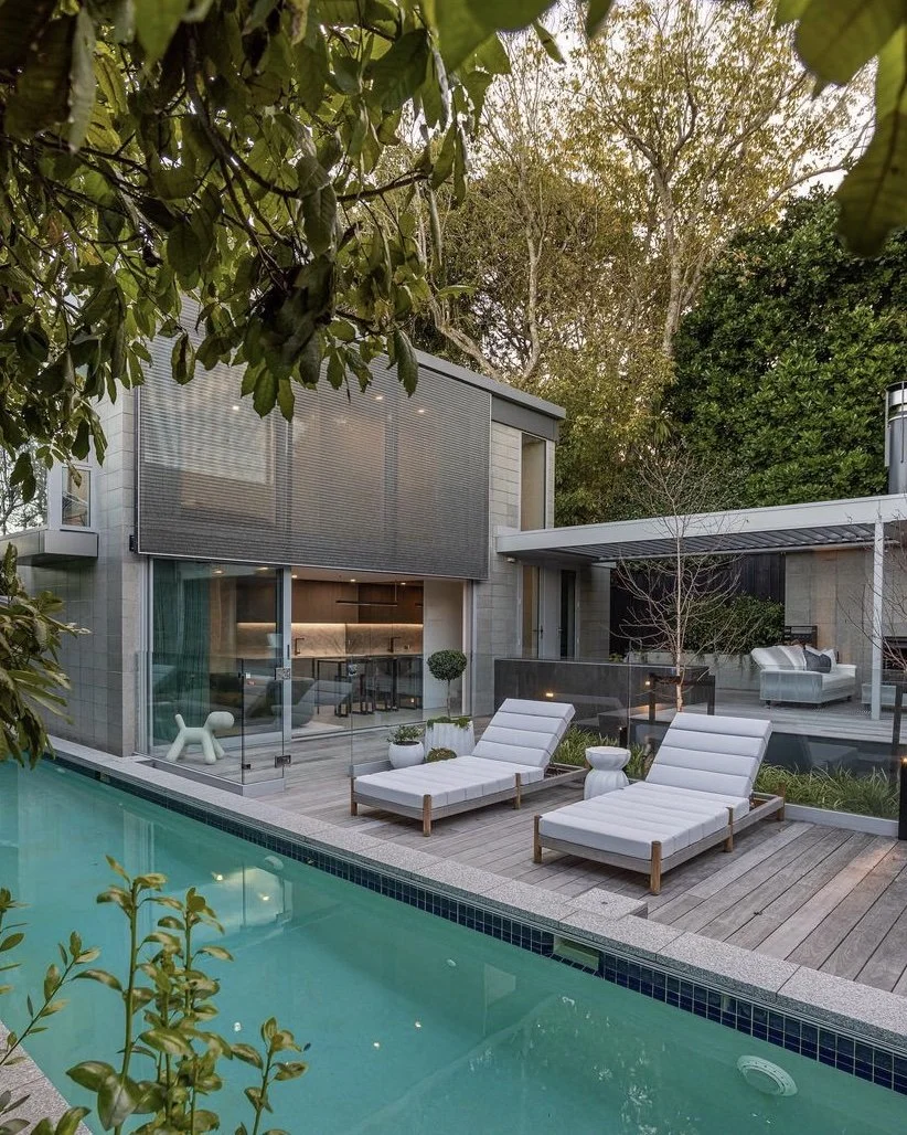 architectural Modern backyard with a swimming pool, two white lounge chairs, plants, a residential house with large glass windows, and outdoor custom patio seating area surrounded by trees.