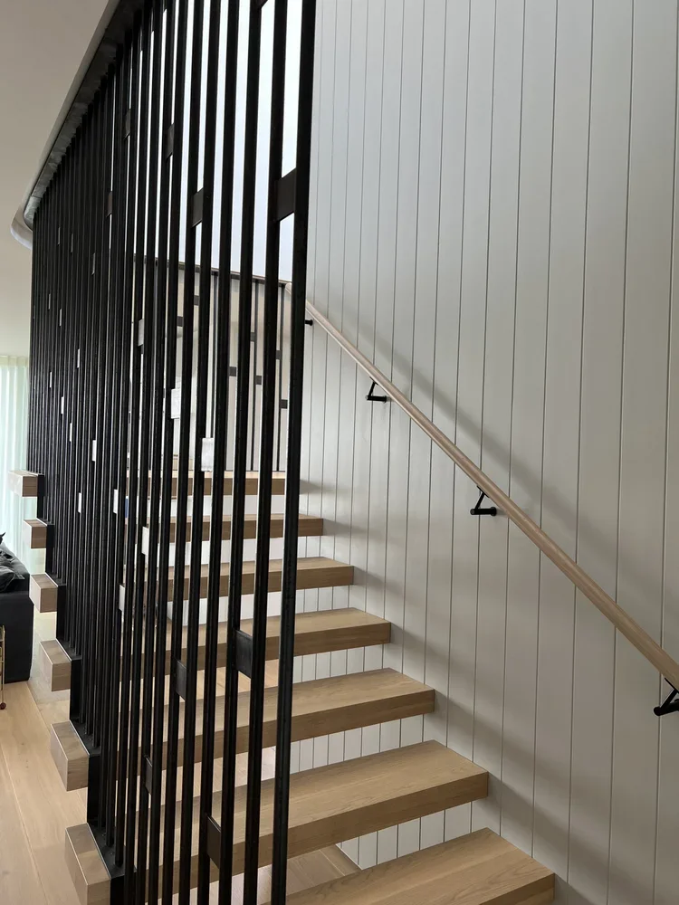 Interior view of a modern staircase with wooden steps, black vertical metal balustrades on the left side, and a wooden handrail on the right side attached to a white paneled wall.
