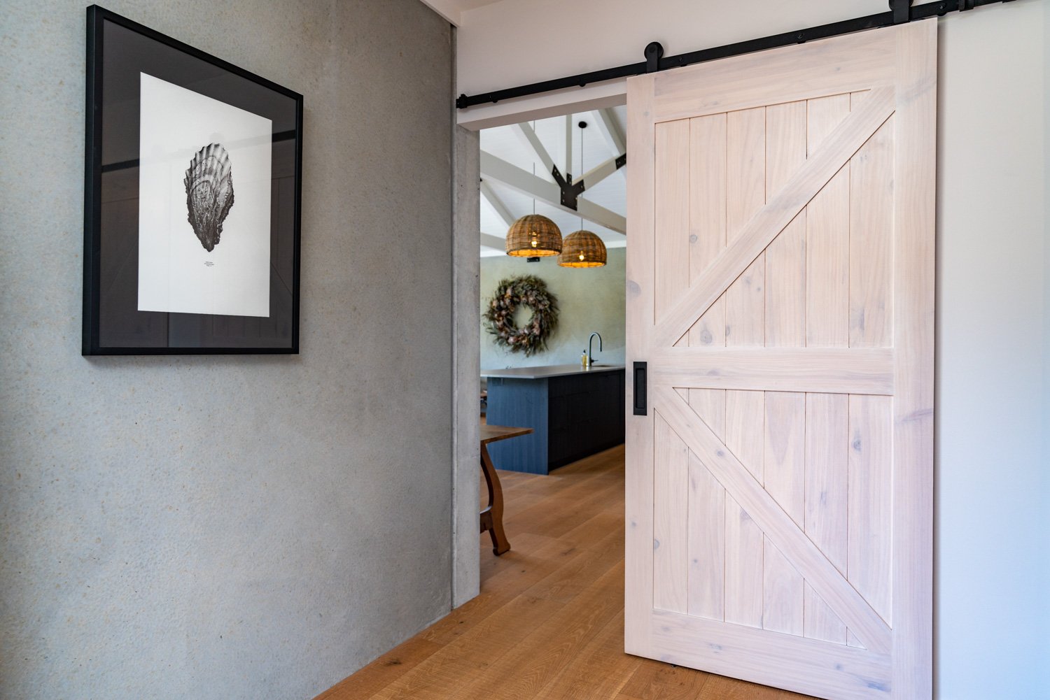 Interior view of a room with a sliding barn door, a framed black and white shell print on the wall, and a glimpse of a kitchen with pendant lights and a wreath hanging on the wall.