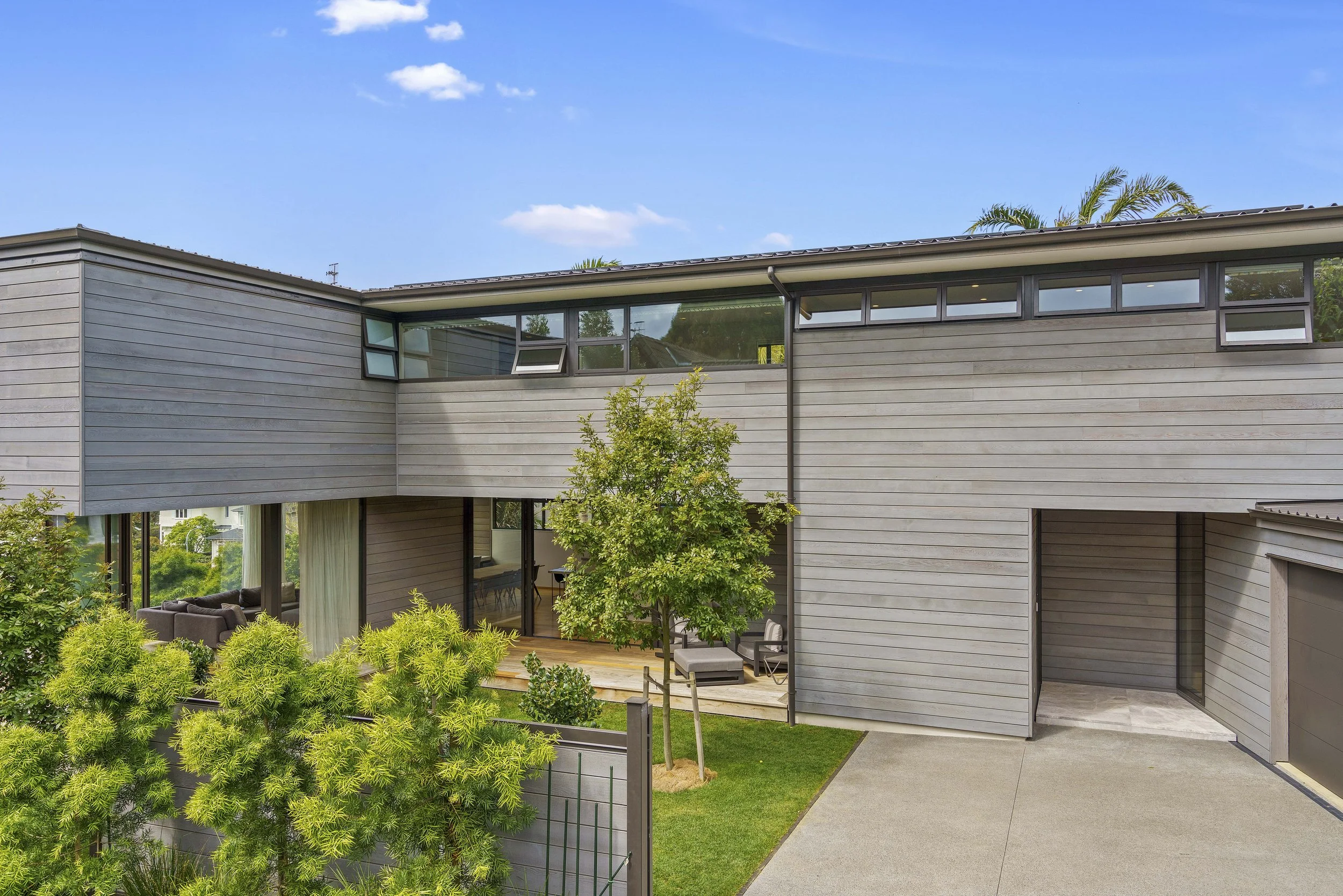 Architectural Modern house with wooden exterior and large windows, surrounded by green trees and a manicured lawn, under a clear blue sky.