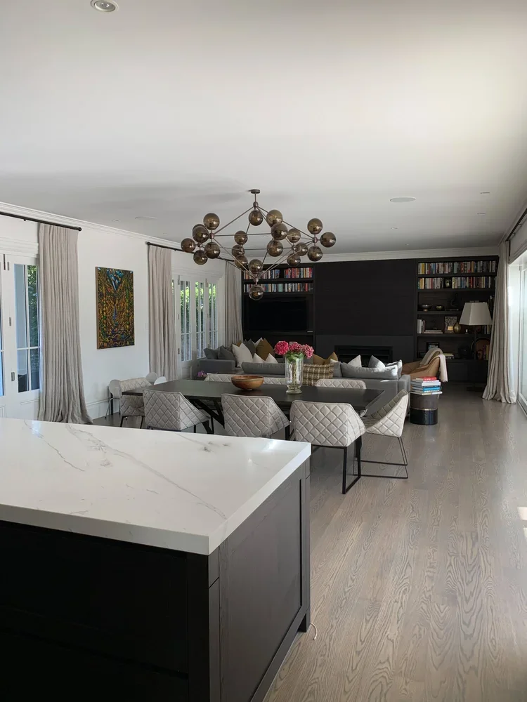 Bright living room with a dining area, gray sofa with pillows, black fireplace, built-in bookshelves, chandelier, artwork on the wall, and large windows with curtains.