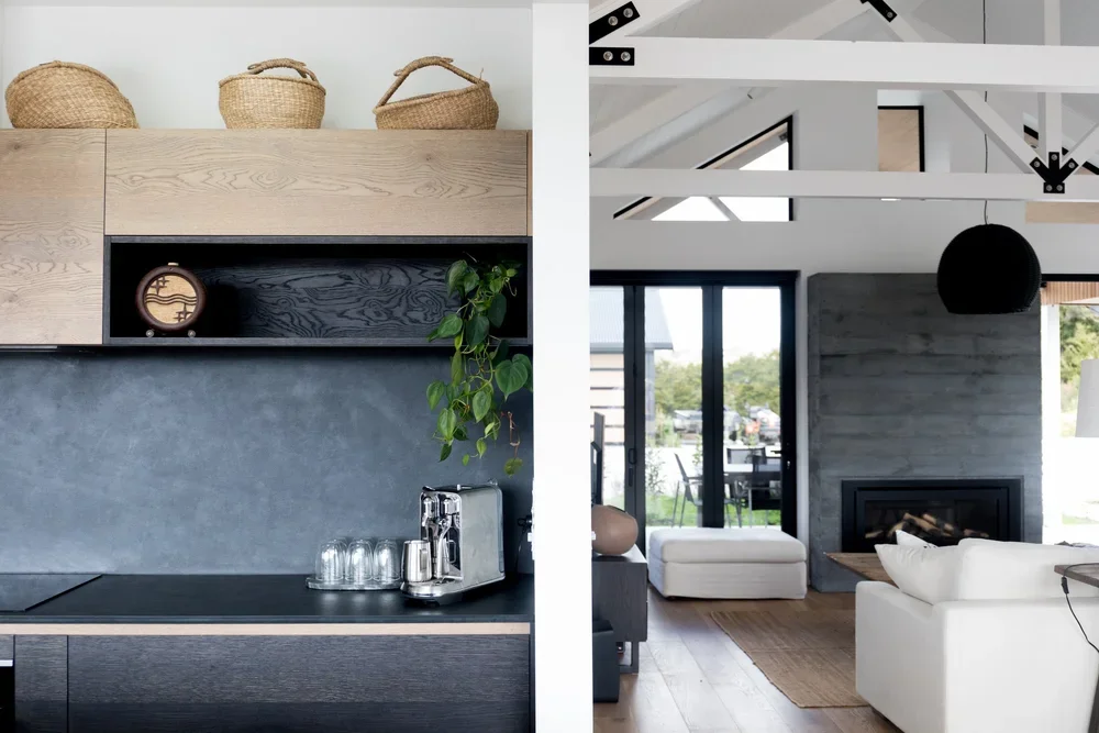 Split view of a modern kitchen on the left with wooden cabinets, open shelving, a coffee machine, and glasses. The living room on the right features a fireplace, large sliding glass doors, white sofa, and vaulted ceiling with exposed beams.