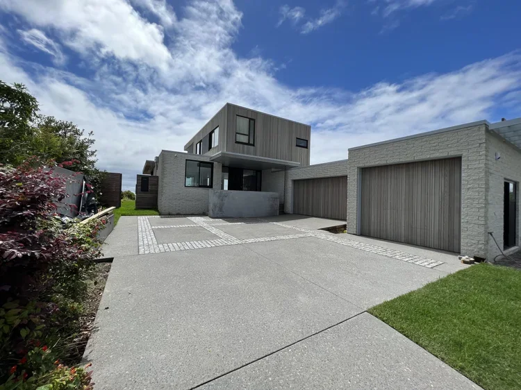 Modern two-story house with gray brick and wood exterior, multiple garage doors, a driveway, and landscaped side yard under a partly cloudy sky.
