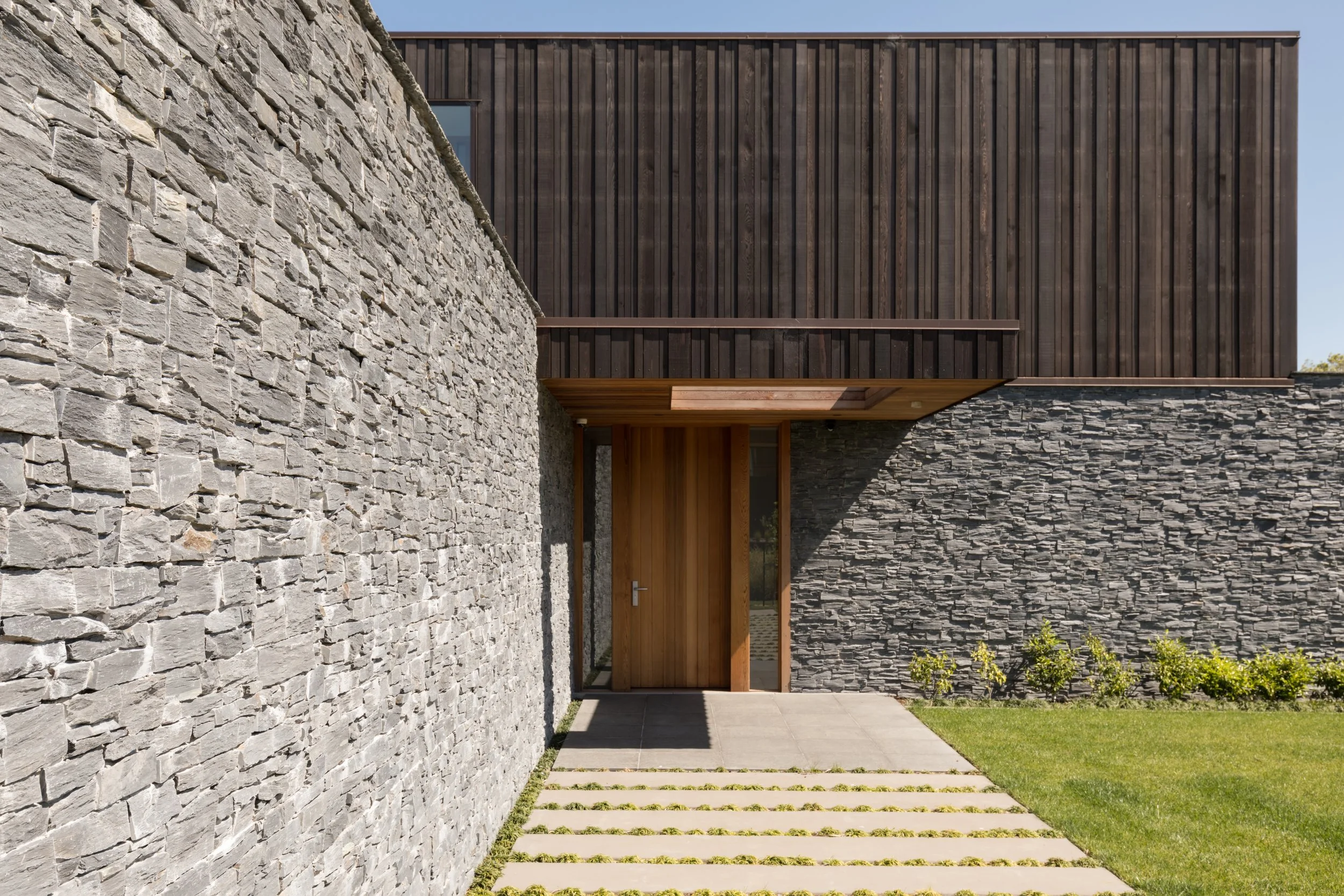 Exterior of a modern architectural palm springs inspired home with a stone wall, wooden paneling, and a pathway with grass and stepping stones.