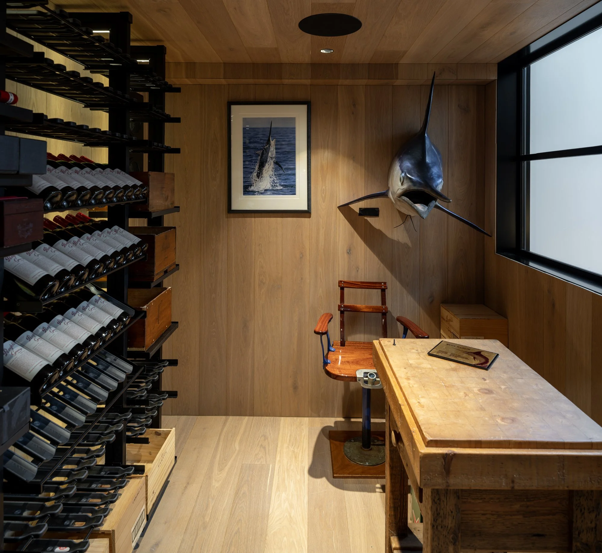 Interior of a custom wine cellar with wooden walls and flooring, wine bottles on black shelves, a framed picture of a fish jumping out of water, and a mounted marlin fish above a small wooden table with chairs.
