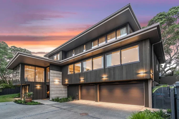 Modern custom multi-story luxury home with expansive windows, dark cladded exterior, and a three-car garage, at sunset.