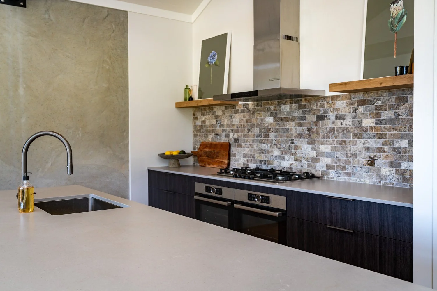 Modern kitchen with beige countertop, stainless steel sink, black cabinetry, gas stove, stone tile backsplash, and wooden shelves.