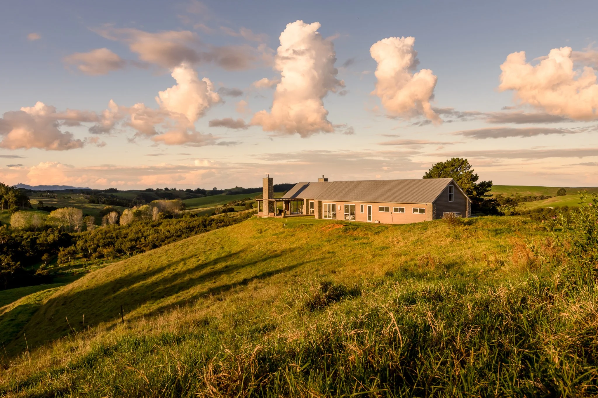 A picturesque custom rural home with a gray roof and large windows is situated on a green hillside, with rolling hills and trees in the background under a sky with scattered clouds at sunset.