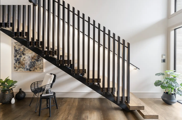 Interior view of a modern stairway with dark wooden steps and black metal railing, situated in a room with wooden flooring, a chair with a cushion, a small table, indoor plants, a framed abstract artwork, and large windows allowing natural light.