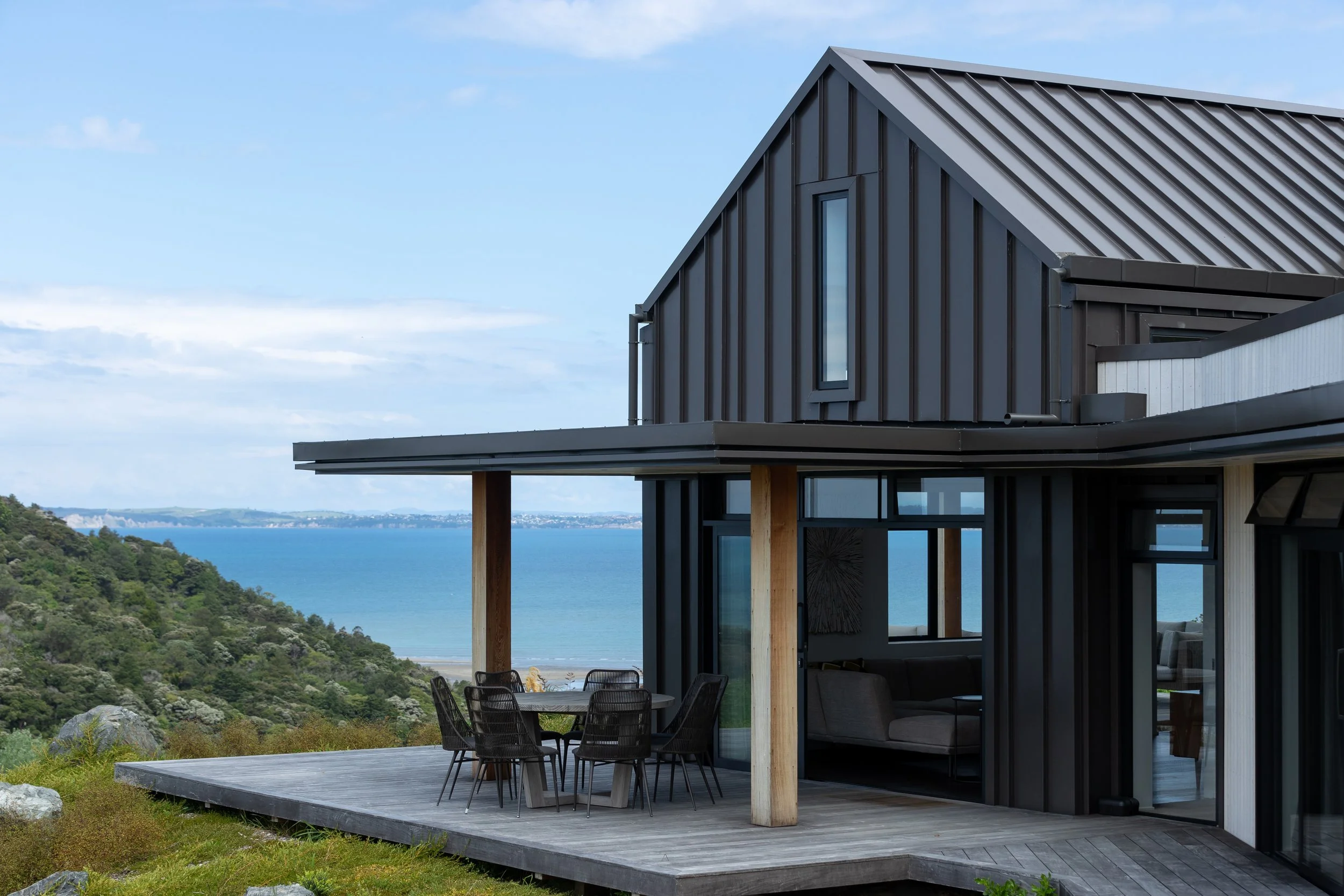 Modern house with black metal siding and large windows, overlooking a scenic landscape with a body of water and green hills, featuring an outdoor deck with black chairs and a dining table.