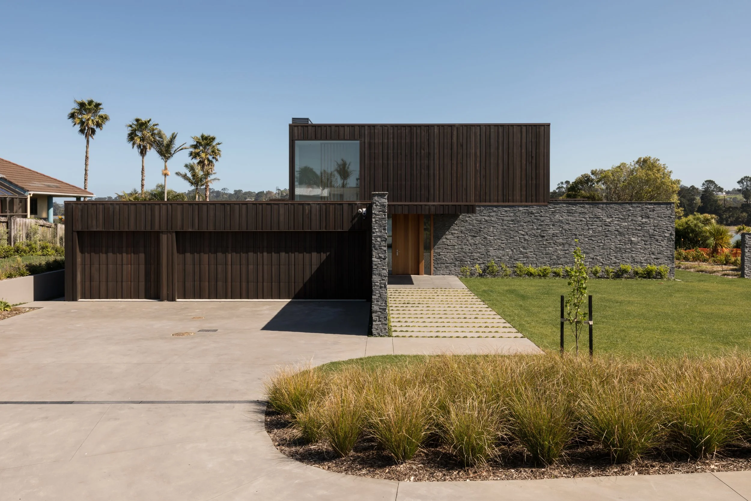 Custom modern house with a flat roof, dark wooden exterior, and stone accents, set in a landscaped yard with palm trees and a driveway.